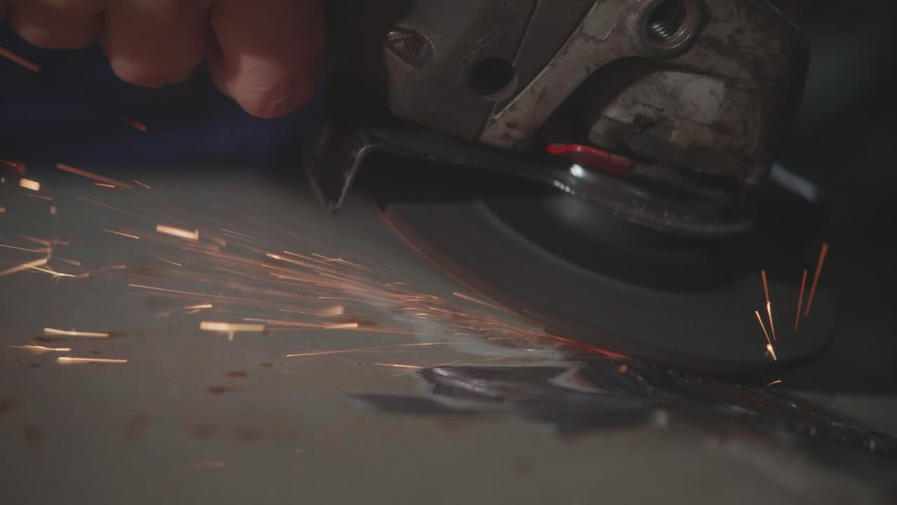 A slow-motion macro shot of a worker using an angle grinder to smooth a weld on sheet metal. Bright sparks fly in a display of industrial metalwork and skilled craftsmanship