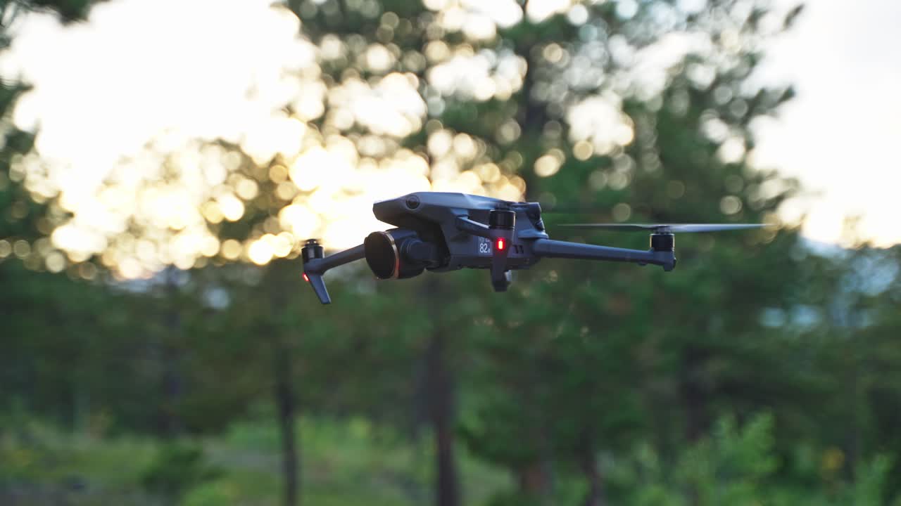 Close-up of a drone hovering steadily in front of trees, captured during soft golden hour lighting in a forested setting, side medium