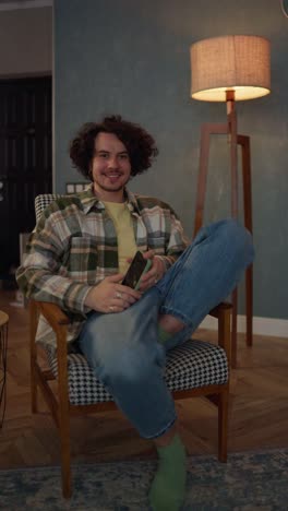 Vertical-video-portraits-on-the-left-of-a-brunette-guy-with-curly-hair-in-a-checkered-shirt-and-jeans-who-sits-on-a-chair-near-a-lamp-with-soft-light-in-a-modern-apartment-at-home