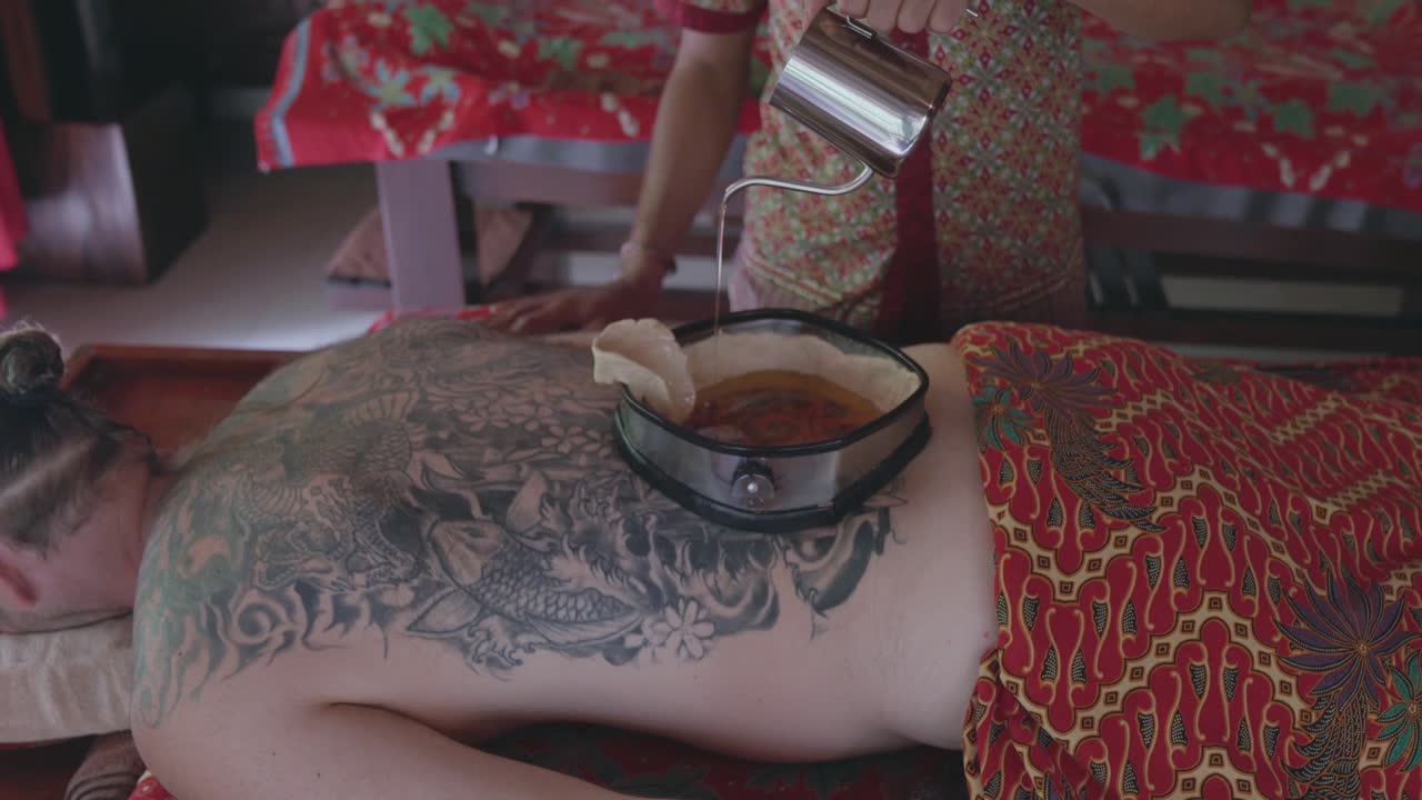 La terapista della spa versa olio caldo in un contenitore rivestito di cera sulla schiena di un paziente maschio che sta ricevendo un trattamento ayurvedico Kati Vasti, poi si allontana