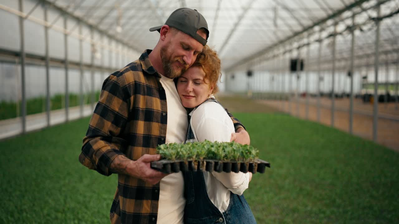 una pareja amorosa de agricultores un tipo rubio y su esposa con el pelo rojo posan en un invernadero entre los brotes de plantas jóvenes en la granja