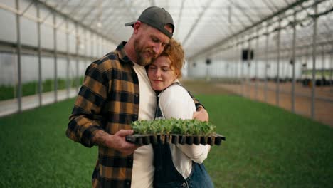 Una-Pareja-De-Agricultores-Enamorados,-Un-Chico-Rubio-Y-Su-Esposa-Pelirroja,-Posan-En-Un-Invernadero-Entre-Los-Brotes-De-Plantas-Jóvenes-En-La-Granja.