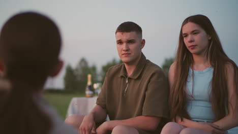 close up rear view of person talking to couple sitting on field as man lifts hand reacting to insects flying around while woman listens attentively capturing peaceful evening interaction outdoors