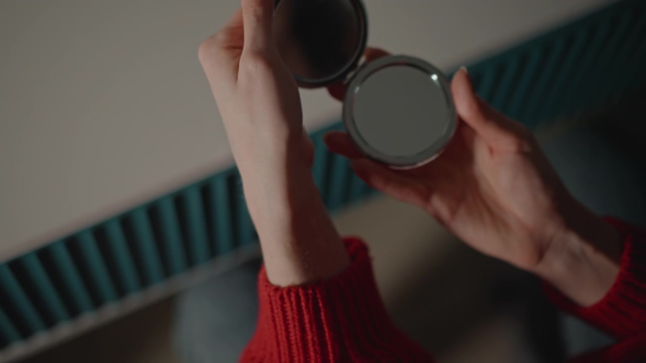 Close-up view of both hands holding open compact mirror over matte tabletop while angling and rotating it, showing manicured nails, red knit sleeve, soft warm light, and calm self-care mood