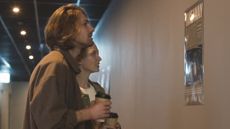 happy couple with a takeaway drink talking together while looking a wall poster at the cinema