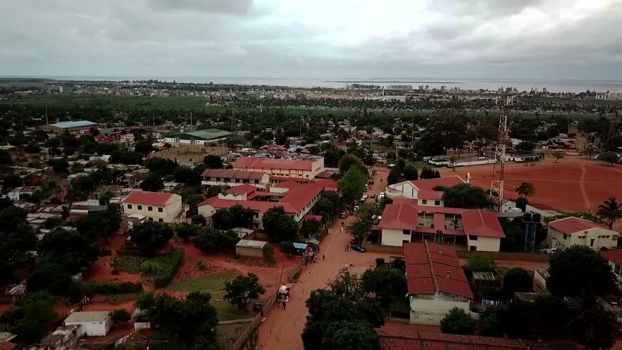 Vista aérea de um bairro suburbano em Maputo, Moçambique, com casas de telhados vermelhos, estradas de terra, árvores espalhadas, e o horizonte distante da cidade e a baía sob um céu nublado.
