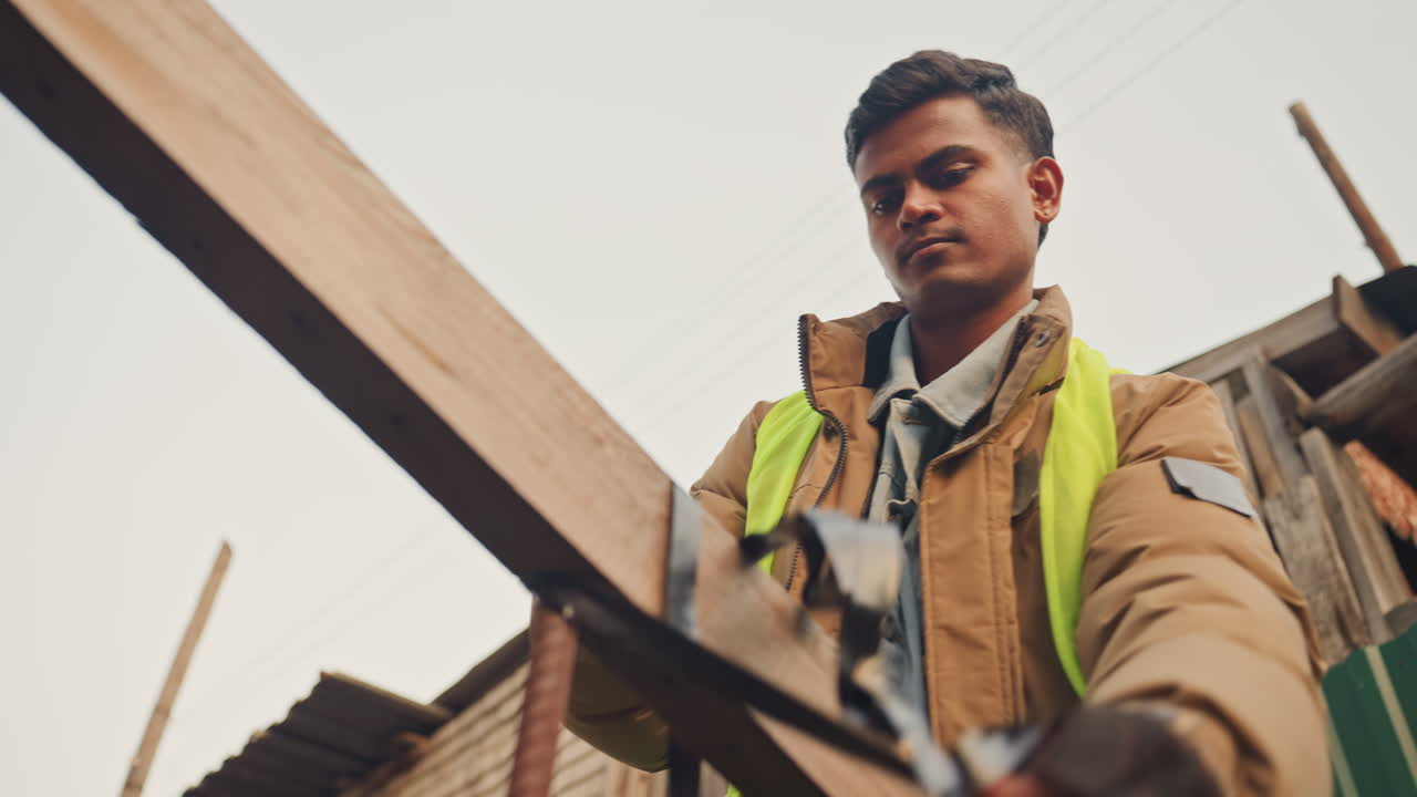 Worker measuring wooden beam for repair and alignment. Asian construction worker in highvisibility vest and gloves uses tape and tools to check timber join, secures hardware and prepares framework