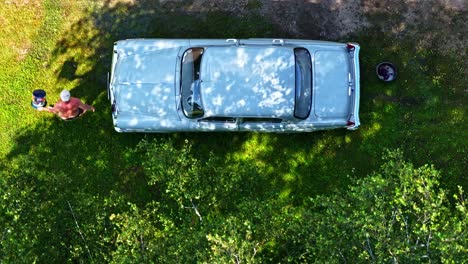 man hand-washing his classic, vintage soviet-era car on a sunny green lawn in the latvian countryside - a top-down aerial slow-motion