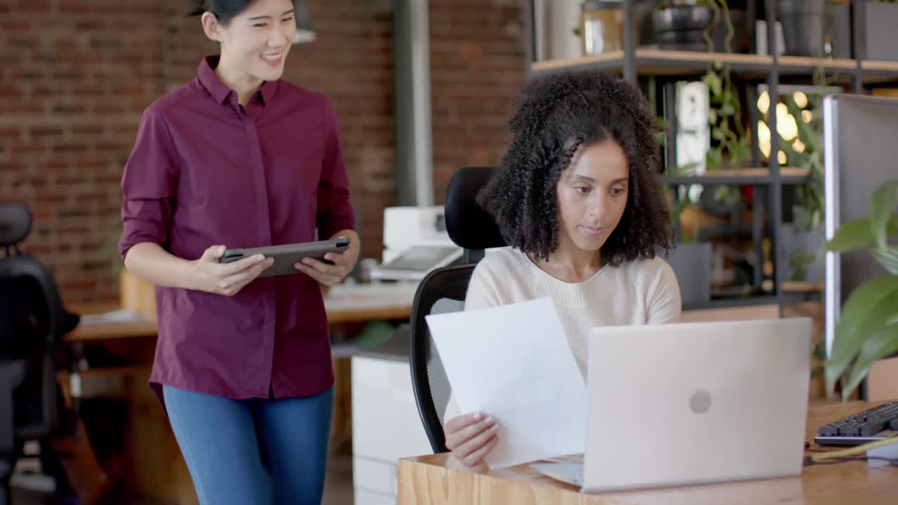 Felici donne d'affari diverse e in abbigliamento casual che usano un tablet, un laptop e parlano in ufficio al rallentatore.