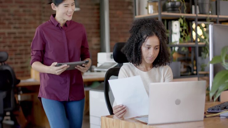 Felici donne d'affari diverse e in abbigliamento casual che usano un tablet, un laptop e parlano in ufficio al rallentatore.