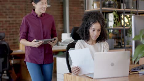 mujeres de negocios casuales felices y diversas usando tabletas, computadoras portátiles y hablando en la oficina en cámara lenta
