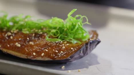 a chef uses is hand and tongs to gently lift a garnished miso glazed eggplant onto a plate for presenting to his guests
