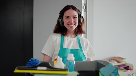 retrato de una feliz chica morena con auriculares inalámbricos negros en una camiseta blanca y un delantal azul que sostiene en sus manos un cuenco de plástico gris con herramientas de limpieza y detergentes en un apartamento moderno