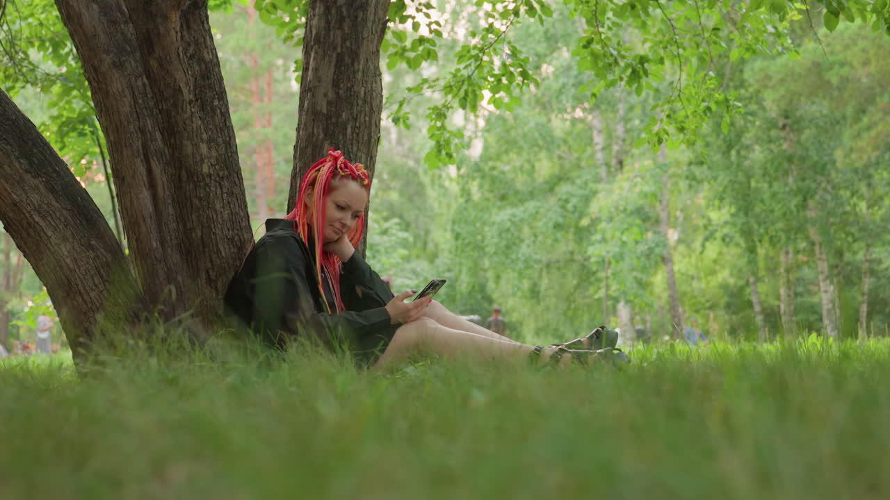 Una persona si rilassa scrivendo un diario sotto un albero, in una scena serena che ritrae una donna immersa in una sessione di scrittura creativa, in un parco tranquillo, con una donna con dreadlocks rossi intenta in una composizione silenziosa.