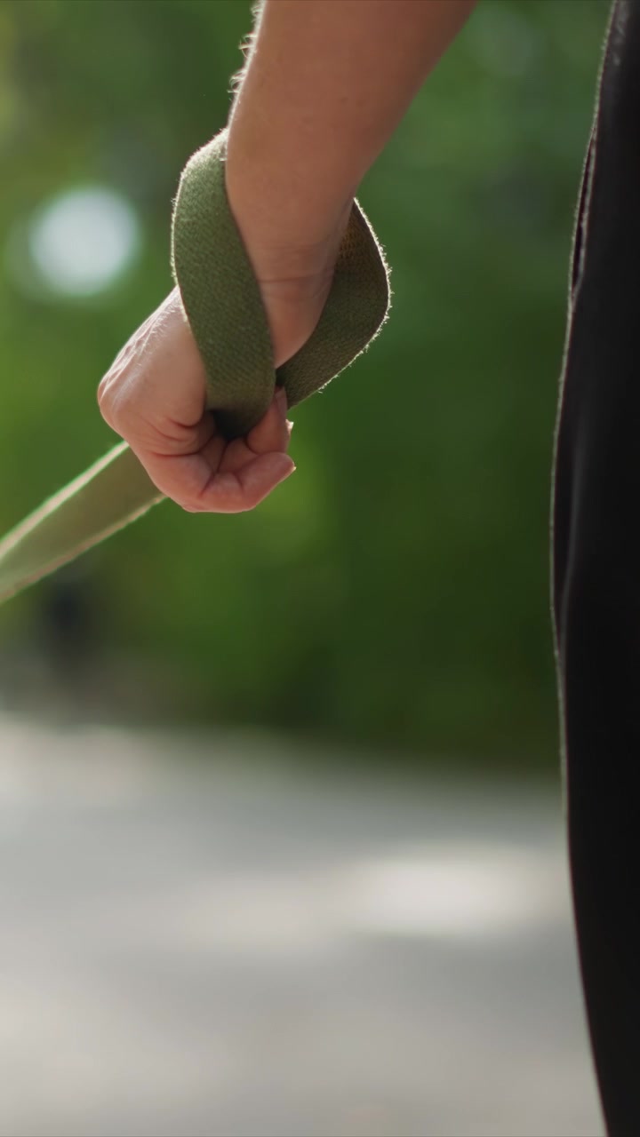 Una persona cammina con una presa visibile su un oggetto collegato. Le mani afferrano saldamente il materiale durante un movimento minimo. Vegetazione sfocata circonda la figura che cammina sotto una forte luce diurna.
