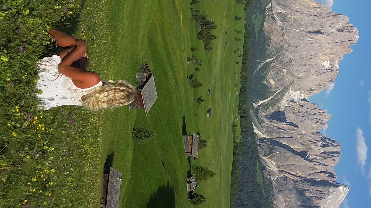 Romantic scene with a beautiful blonde girl sitting on a meadow with green grass and flowers in Alpe di Siusi, Dolomite Mountains, Italy