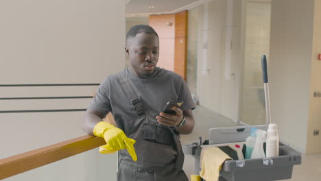 cleaning man wearing gloves leaning on the stair railing while looking at his smartphone next to the cleaning cart