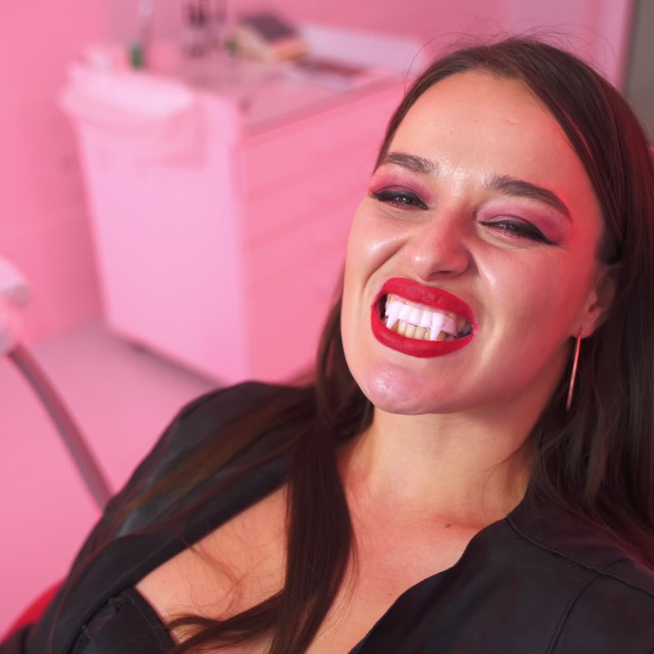 Dark-haired woman in dentist's chair looks at her vampire teeth in the mirror. Lady with bright makeup is happy about her reflection. Close up