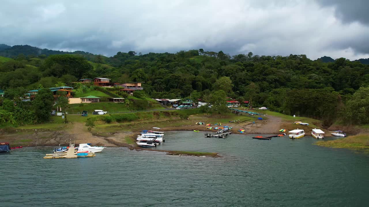 Drone approaches a small lakeside village near Lake Arenal from over the water, moving inland toward the harbour and brightly coloured rooftops