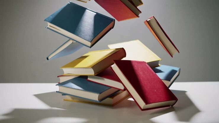 La pila di base si sposta, facendo cadere libri con copertina rigida su un tavolo bianco, proiettando ombre nette.