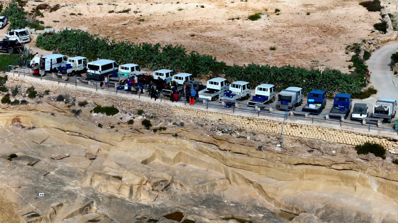 Malta'da parlak güneşli bir yaz gününde, kireçtaşı uçurum kenarında park edilmiş vintage beyaz ve mavi kamyonetler ve minibüsler sırası; engebeli kurak kıyı manzarasının yüksek açılı geniş bir çekimiyle.