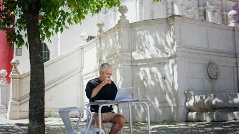mature businessman drinking coffee at street cafe. focused senior working laptop