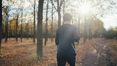 Rückansicht-Eines-Mannes-Mit-Lockigem-Haar-In-Einer-Schwarzen-Sportuniform,-Der-Beim-Morgendlichen-Joggen-In-Einem-Herbstmorgenwald-Bei-Strahlender-Sonne-Auf-Einem-Erdweg-Läuft