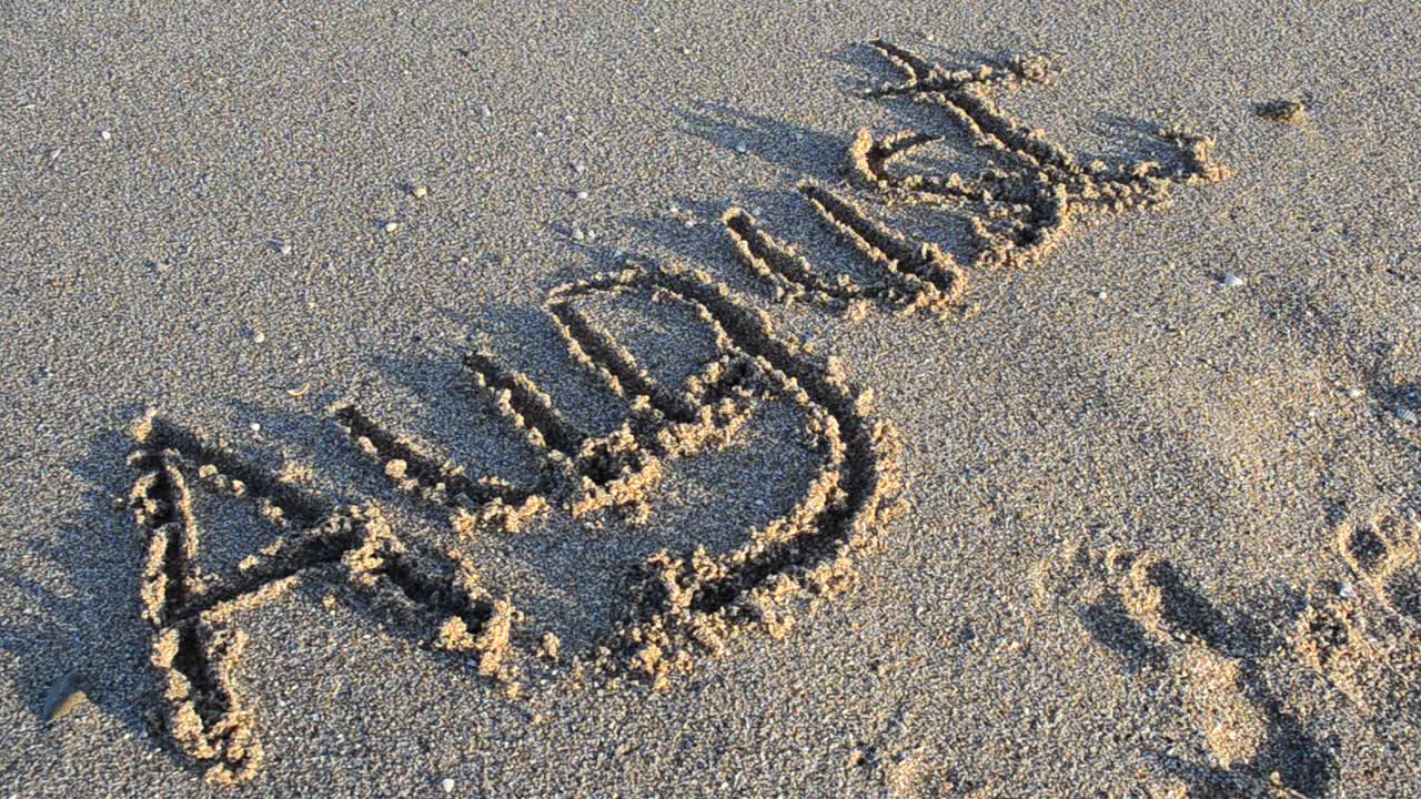 Scritta 'agosto' sulla sabbia. La scritta viene spazzata via da un'onda del mare. Riprese sulla spiaggia.