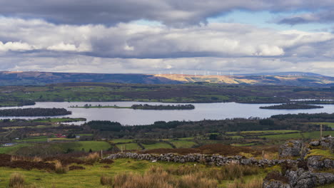 Timelapse-of-rural-nature-farmland-with-stonewall-in-the-foreground-and-wind-turbines-in-hills-and-lake-in-distance-during-sunny-cloudy-day-viewed-from-Carrowkeel-in-county-Sligo-in-Ireland