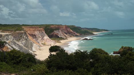 Tilt-up-shot-revealing-the-stunning-Northeast-Brazil-coastline-in-Paraiba-with-huge-colorful-clay-cliffs,-golden-sand,-and-crystal-clear-turquoise-water-on-a-sunny-summer-day-with-a-breeze