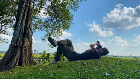 Relaxed-young-white-man-taking-pictures-of-sky-lying-on-grass-next-to-tree