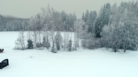 aerial flight during falling snowflakes during white winter day in wilderness - wooden house and barell sauna in nature