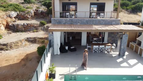 mujer delgada de cabello oscuro en vestido de verano toma un sorbo de un vaso y sale de una casa a una piscina al aire libre - drone vuela alto fuera de la villa en grecia creta con palmeras