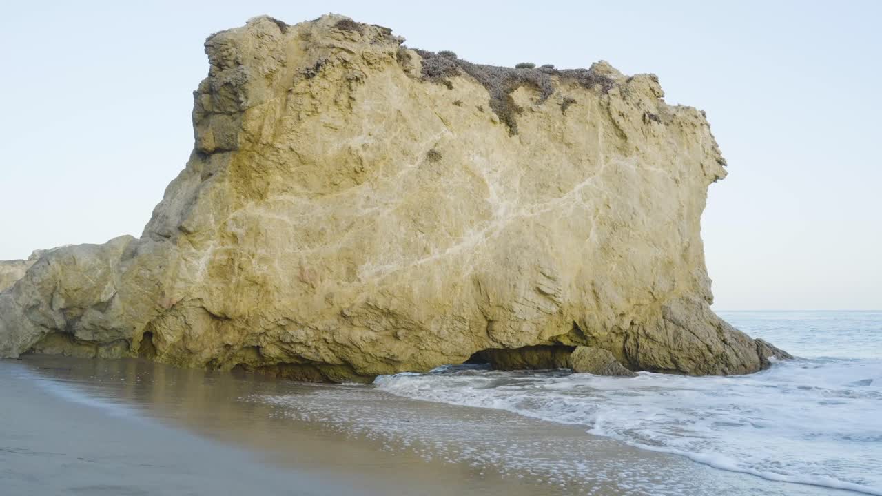 Ocean waves crash gently at the base of a large coastal rock formation during daylight on a quiet beach