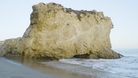 ocean waves crash gently at the base of a large coastal rock formation during daylight on a quiet beach