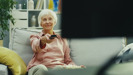 portrait shot of the old woman sitting on the couch in the living room, turning on tv with a remote control and laughing while watching tv