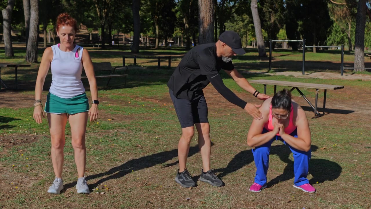 Mandlig fitnesstræner, der retter på form og motiverer to kvinder, mens de laver squats med kropsvægt i en udendørs park en solrig dag