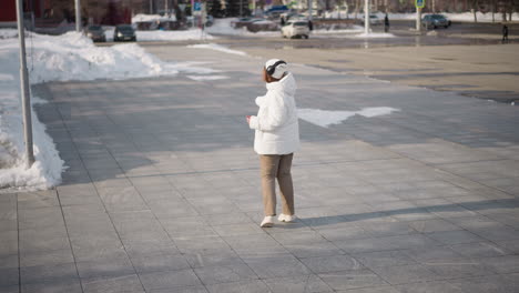 young student wearing white winter coat, headset and sunglasses performs energetic tap dance on tiled plaza under bright sunlight, snowbanks at perimeter, busy road and trees in background