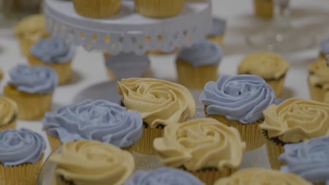 Stockclip-Zum-Hochzeitstag-Mit-Nicht-Enden-Wollenden-Cupcakes,-Blauer-Und-Gelber-Zuckerguss-Mit-Goldener-Cupcake-Papierfolie-Auf-Weißem-Tisch-Und-Tortenständern,-Während-Die-Kamera-Langsam-Nach-Oben-Schwenkt