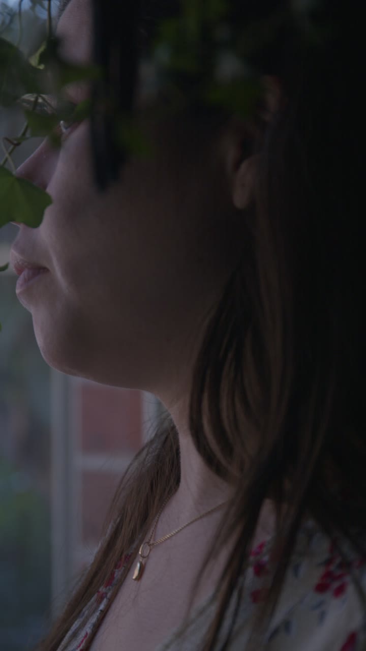 A pensive woman gazes out a window in soft focus. Dim lighting and blurred details create an introspective mood. Captured in vertical video format, perfect for social media