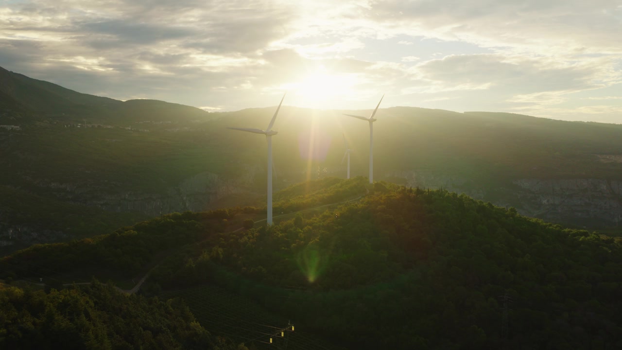 Three wind turbines stand silhouetted atop green hillside crest in Italy, blazing golden sunrise glows behind layered misty mountains above small distant lakeside village. Aerial drone view.