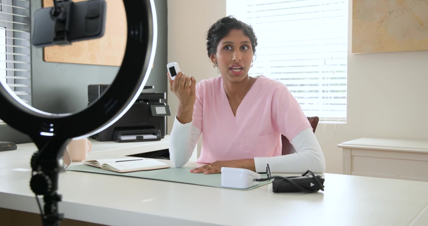 Donna afroamericana in camice che mostra il telefono alla telecamera alla scrivania con una ring light che illumina il dispositivo.