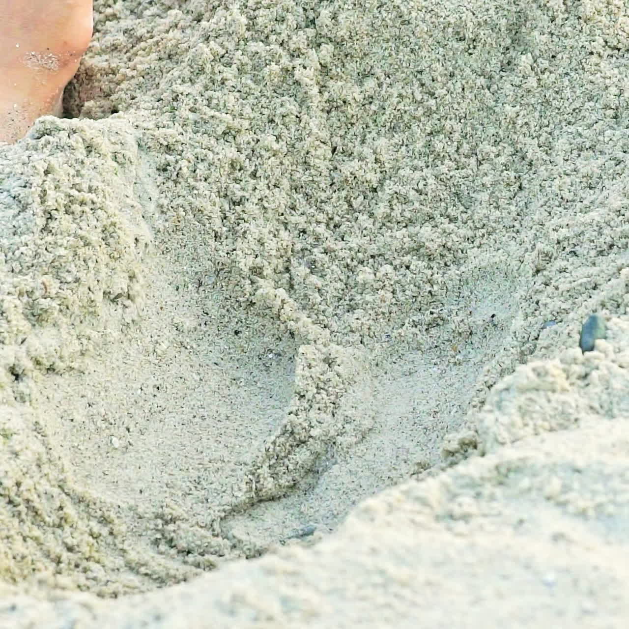 A kid gets up in a sandy pit with his legs to measure it and continues to expand its length and width. Close-up.
