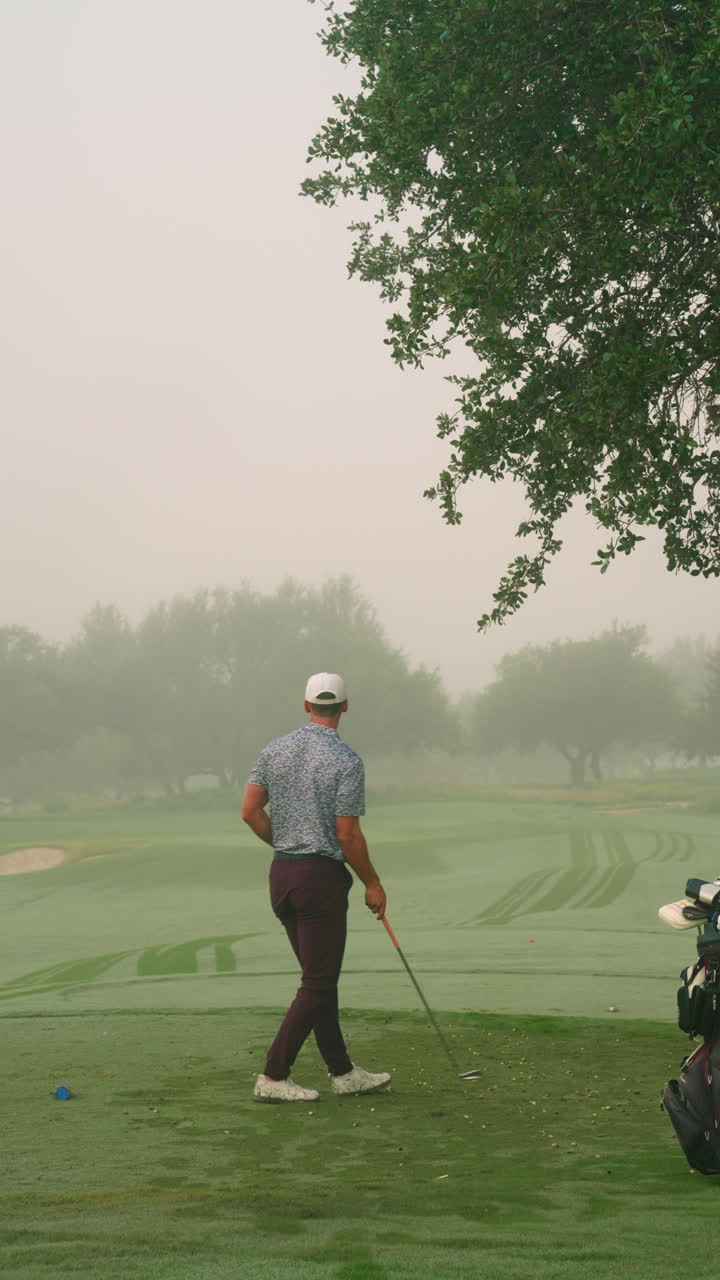 Inquadratura verticale da dietro e angolata di un golfista che colpisce la pallina con un ferro su un tee in una mattinata nebbiosa, con il segnaposto visibile mentre conclude il colpo con una fluida rotazione del bastone accanto alla sacca da golf