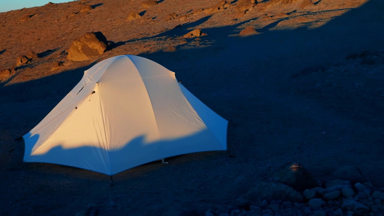 Una tenda a cupola bianca è piantata su ghiaia grossolana e massi sparsi su un pendio desertico brullo, illuminata da una calda luce solare radente che proietta lunghe ombre su