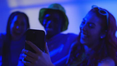 Foto-De-Estudio-De-Un-Grupo-De-Amigos-De-La-Generación-Z-Sentados-En-Un-Sofá-Posando-Para-Un-Selfie-En-Un-Teléfono-Móvil-Por-La-Noche-Con-Una-Luz-Intermitente-4