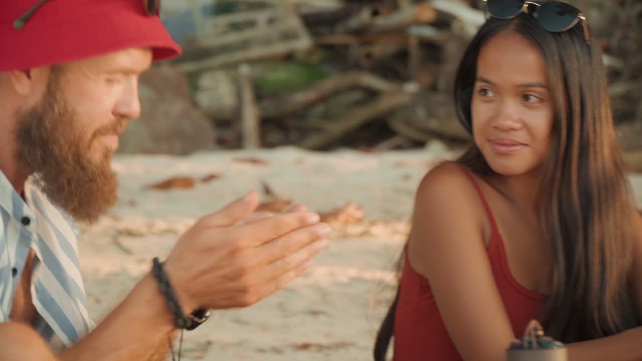Coppia che condivide il tè in spiaggia, donna in costume da bagno rosso con trecce e occhiali da sole, uomo barbuto con cappello rosso che scambia una tazza dal thermos, sfondo di legni levigati dal mare e litorale, luce dorata del tramonto, atmosfera soffusa.