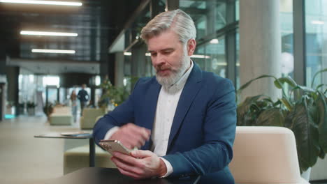 mature businessman using smartphone becomes shocked sudden lottery results in office hotel lobby