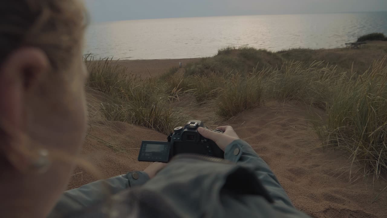 Panning over the shoulder of a female squatting down on sandy dunes holding a camera filming the sunrise over the ocean on an overcast morning surrounded by lush green grass