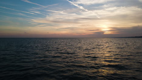 Low-flying-aerial-dolly-above-ripples-of-ocean-water-as-sun-sets-behind-clouds